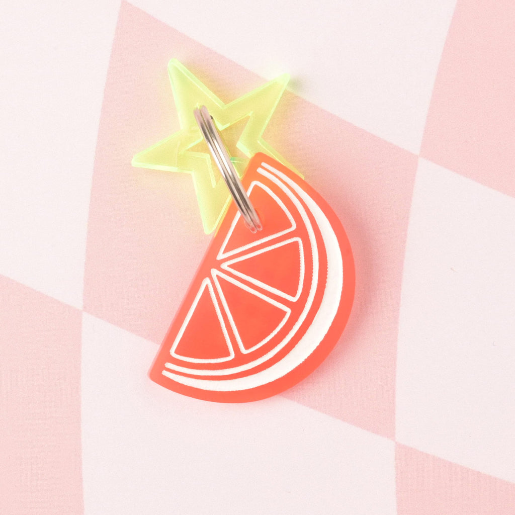 A layered acrylic charm combination: a coral orange slice charm with white detailing layered over and attached to a fluorescent neon yellow star charm. The charms are on a pink and white checkered background.
