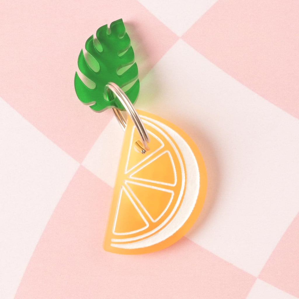 A layered acrylic charm combination: a bright lemon slice charm with white detailing layered beneath and attached to a green tropical Monstera leaf charm. The charms are on a pink and white checkered background.