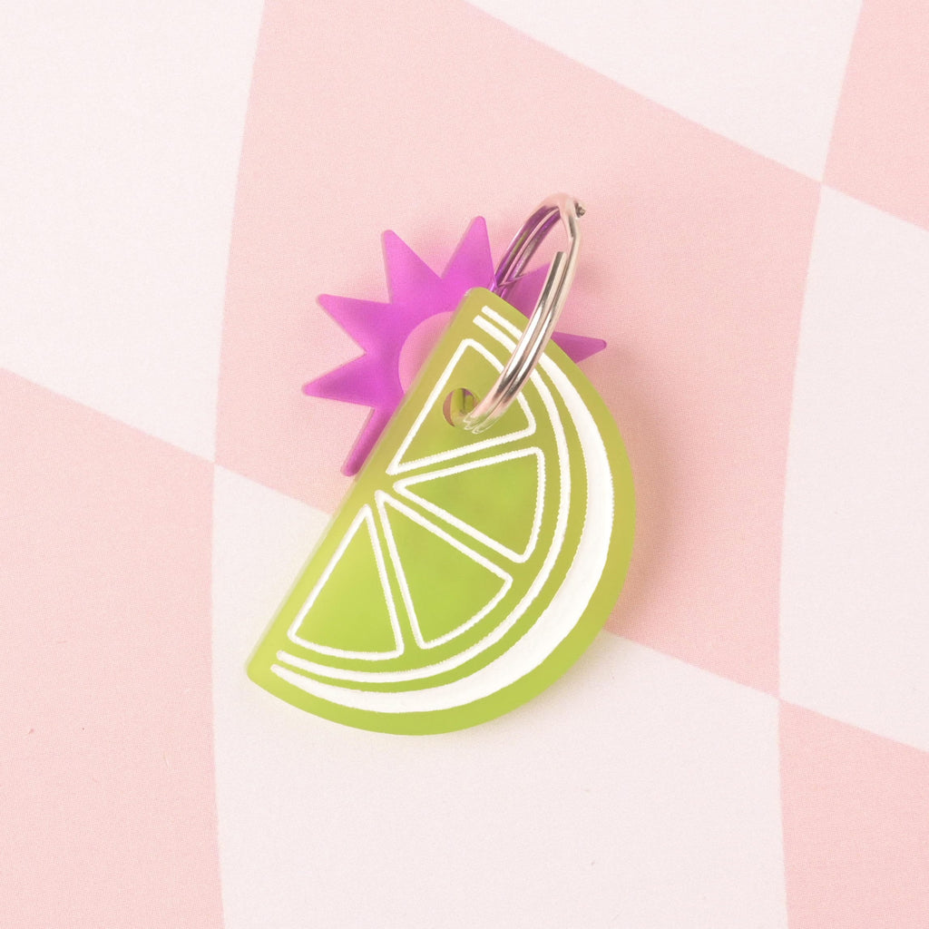 A layered acrylic charm combination: a neon green lime slice charm with white detailing layered over and attached to a translucent pink sunburst charm. The charms are on a pink and white checkered background.