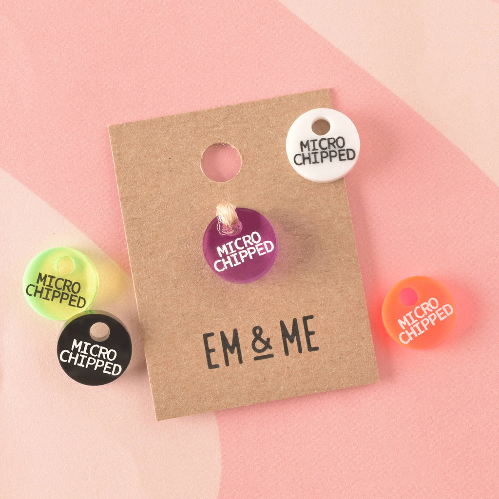 A collection of five small, circular acrylic charms, all engraved with "MICROCHIPPED" in white or black text, and displayed in various colors: white, black, purple, neon yellow-green, and orange. One charm is secured to a branded kraft card.