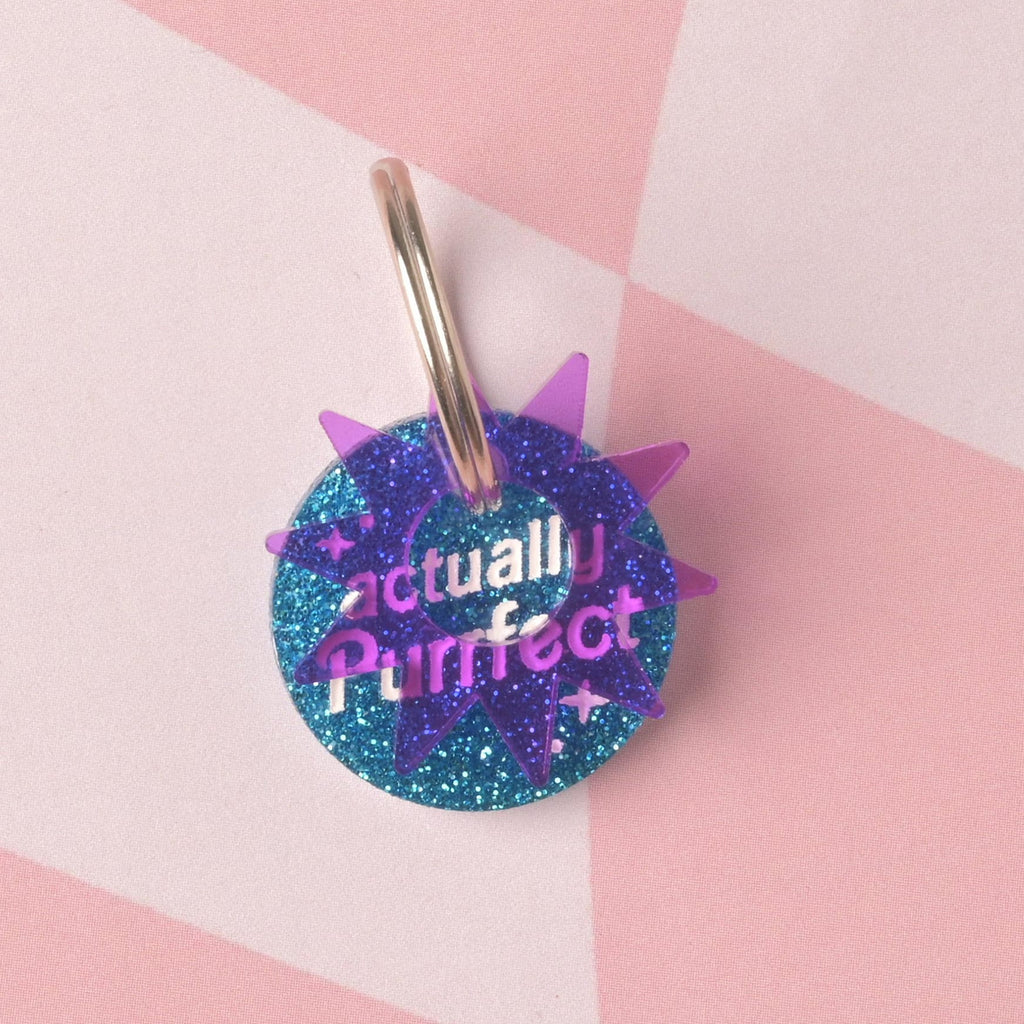 A round acrylic tag made from vibrant blue glitter, engraved with the purple slogan "actually Purrfect". The tag is accented with a translucent purple sunburst charm and shown with a silver ring.
