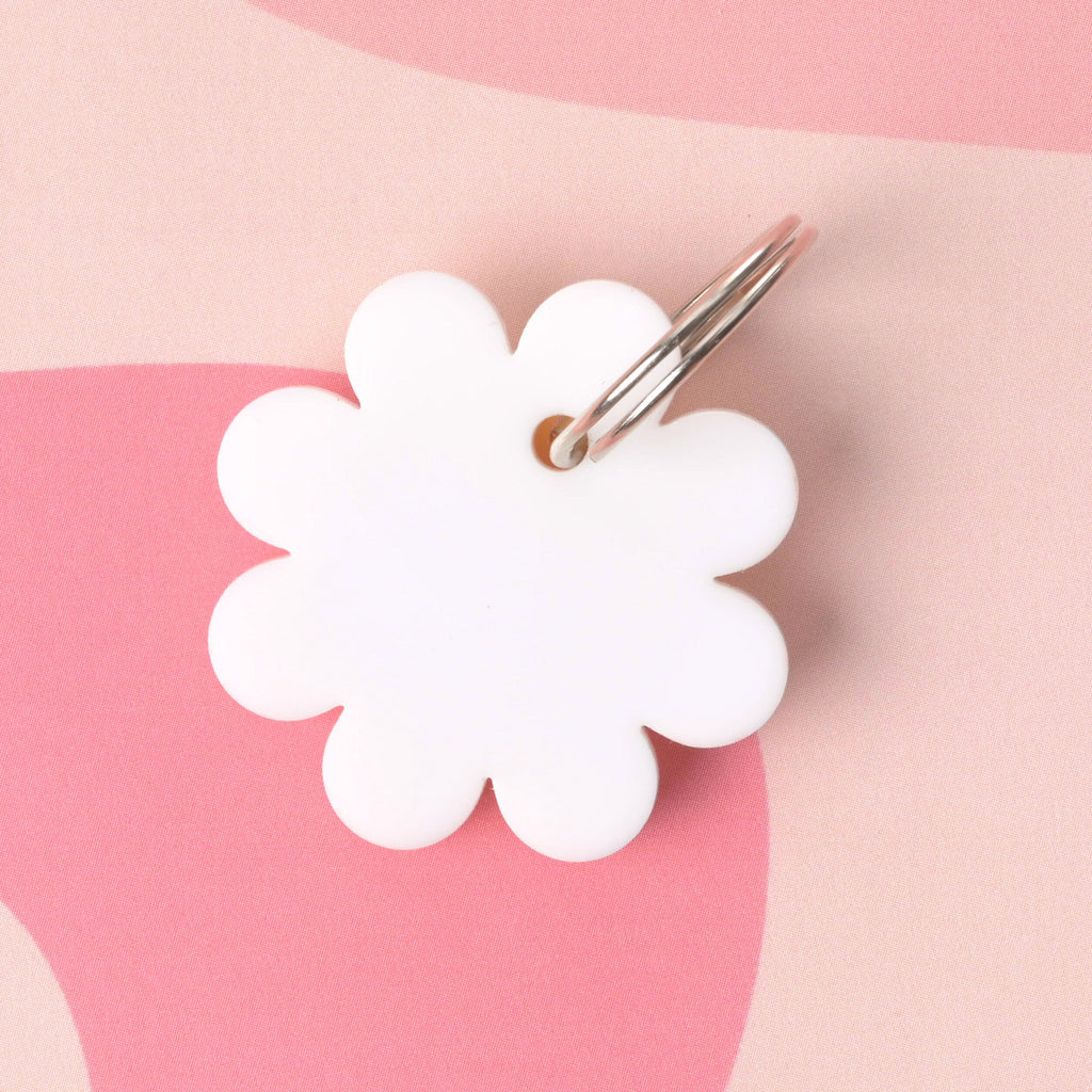 White acrylic flower-shaped pet tag shown from the back with a metal key ring attached, placed on a pink abstract background
