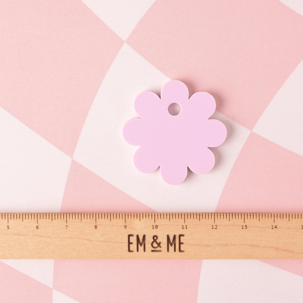 Back view of a lavender daisy-shaped pet ID tag shown next to a wooden EM & ME ruler for size reference, on a pink checkered background