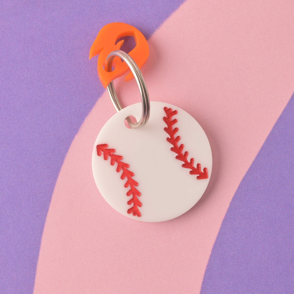 A close-up, overhead view of the front of a white circular acrylic pet ID tag designed like a baseball with red stitching. An orange flame charm is attached to the silver split ring. The tag is displayed on a purple and pink striped background. Baseball Pet Tag, Personalized Pet tag, Cat or Dog ID Tag, summer ball tag
