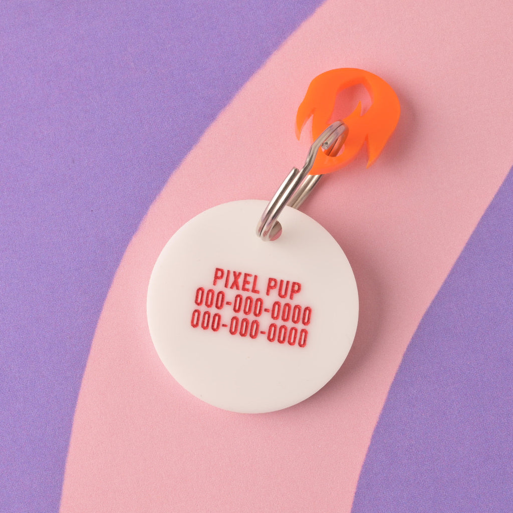 A close-up of the back of a round white acrylic pet ID tag, engraved with the pet's name 'PIXEL PUP' and three lines of placeholder phone numbers in red text. An orange flame charm is attached to the silver split ring. The tag is displayed on a purple and pink striped background. Baseball Pet Tag, Personalized Pet tag, Cat or Dog ID Tag, summer ball tag