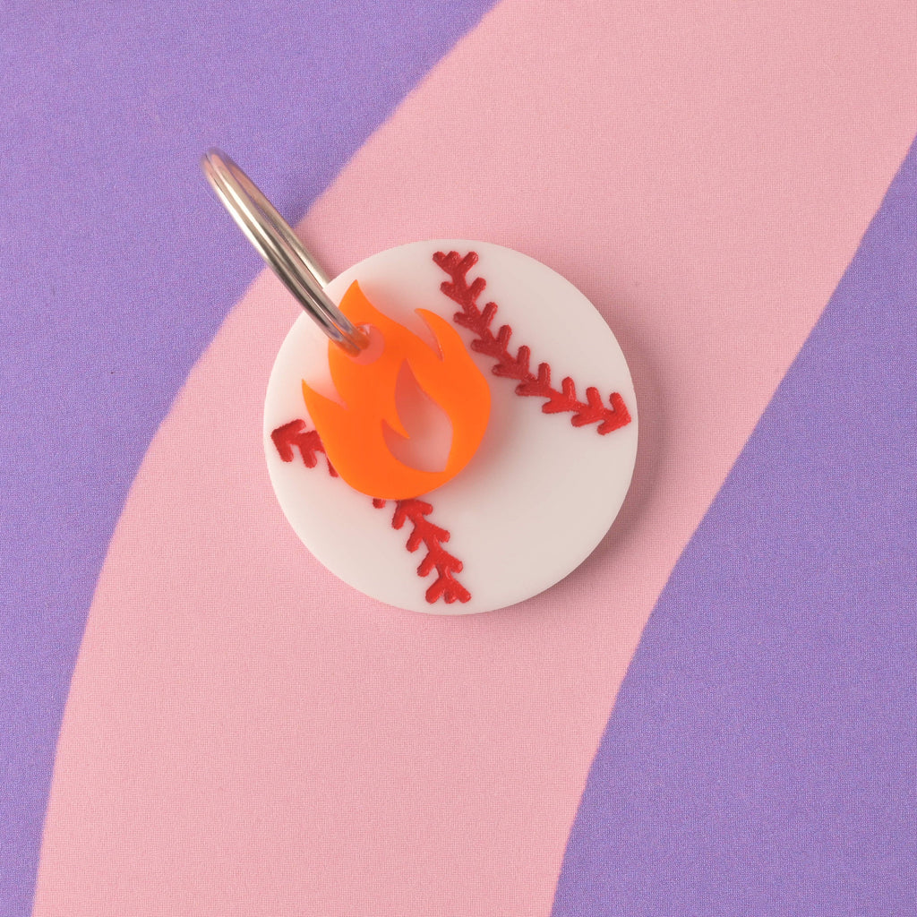 A close-up of the front of a white circular acrylic pet ID tag designed like a baseball with red stitching. An orange flame charm is attached to the silver split ring. The tag is displayed on a purple and pink striped background. Baseball Pet Tag, Personalized Pet tag, Cat or Dog ID Tag, summer ball tag