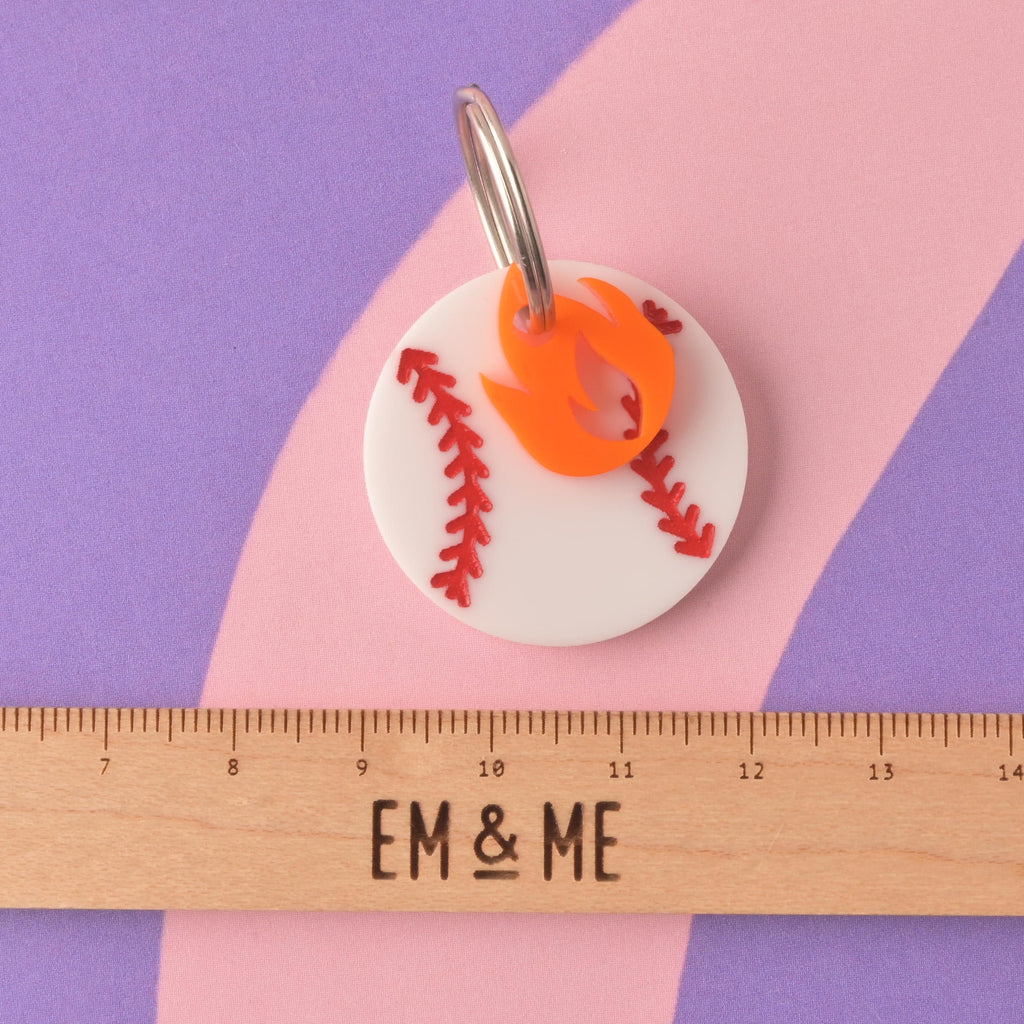 A close-up of a white circular acrylic pet ID tag designed like a baseball with red stitching. An orange flame charm is attached to the silver split ring. The tag is displayed above a wooden ruler marked with the brand EM & ME, on a purple and pink striped background. Baseball Pet Tag, Personalized Pet tag, Cat or Dog ID Tag, summer ball tag