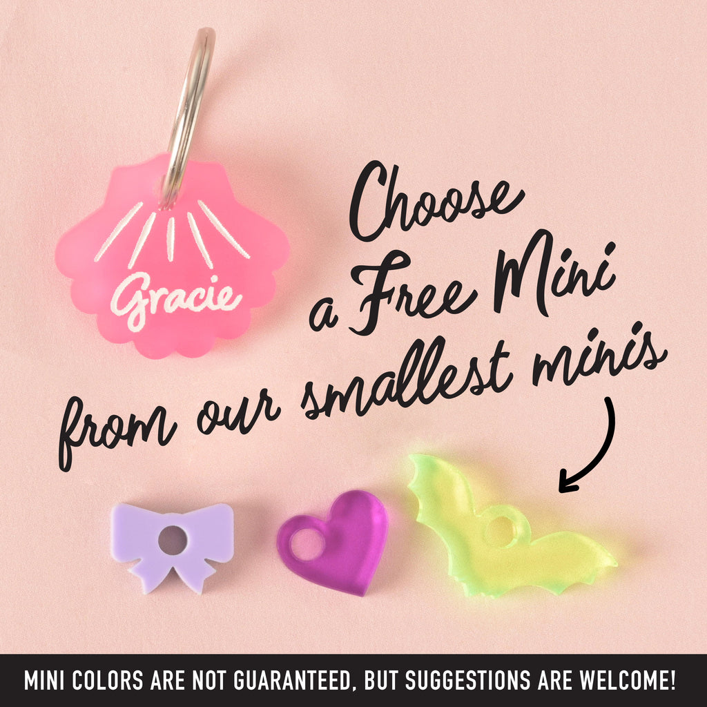 A pink seashell-shaped small tiny pet tag that says "Gracie." The image also shows three small charms—a purple bow, a pink heart, and a translucent yellow bat. The text "Choose a Free Mini from our smallest minis" and "MINI COLORS ARE NOT GUARANTEED, BUT SUGGESTIONS ARE WELCOME!" is visible. The items are displayed on a solid pink background.