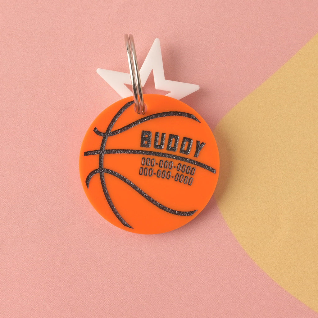 Close-up of a circular orange acrylic pet ID tag designed like a basketball, personalized with the name BUDDY and phone numbers. It is attached to a white star charm and a silver ring, displayed on a pink and yellow background. Basketball, March Madness, Air Bud Personalized Pet tag, Cat or Dog ID Tag