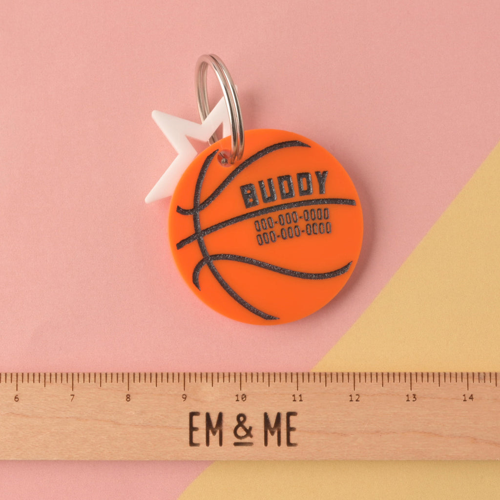 A close-up of a circular orange acrylic pet ID tag designed like a basketball, personalized with the name BUDDY and phone numbers. It is attached to a white star charm and a silver ring, displayed above an EM & ME ruler on a pink and yellow background. Basketball, March Madness, Air Bud Personalized Pet tag, Cat or Dog ID Tag