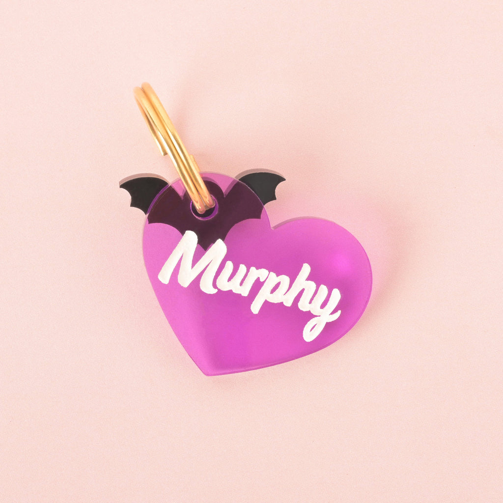 A close-up product photo of a purple, translucent heart-shaped pet tag with small black bat wings attached to the top. The tag is engraved with the name "Murphy" and is shown with a gold split key ring on a pink background.