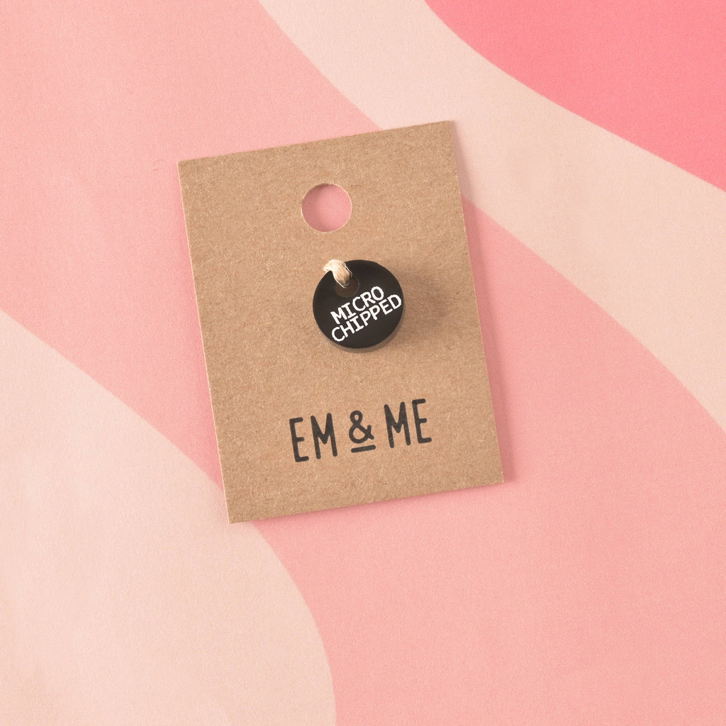 A small, circular, opaque black acrylic charm engraved with "MICROCHIPPED" in white text. The charm is secured with twine to a branded 'EM & ME' kraft card.