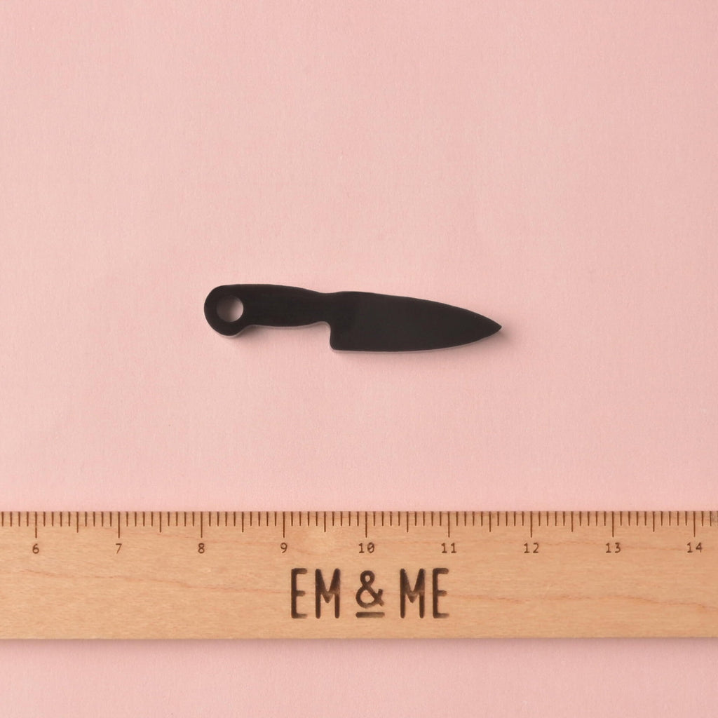 A small, opaque black acrylic mini charm shaped like a chef's knife. The charm is shown above a wooden ruler with the 'EM & ME' logo to indicate its size (approximately 3cm long).