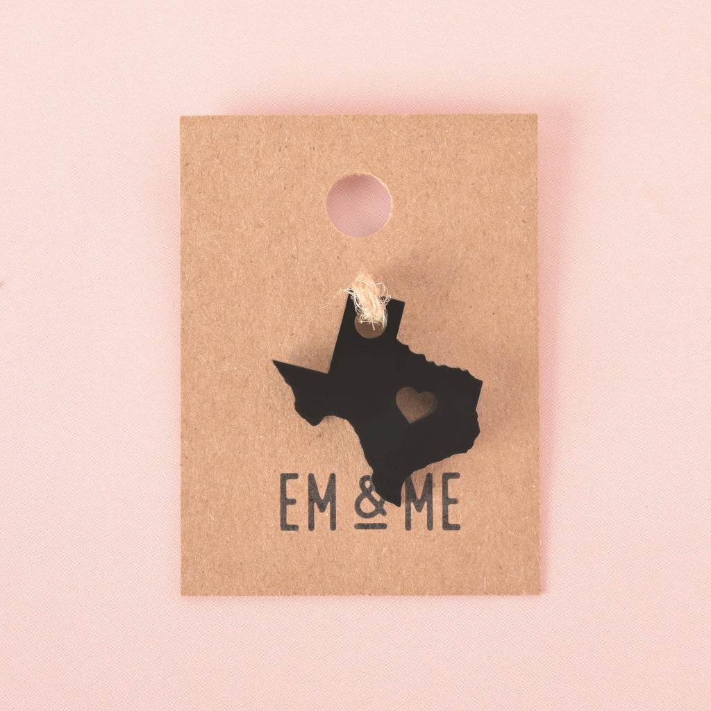 Black acrylic Texas state charm with heart cutout, displayed on kraft paper backing with Em & Me Studio branding, set against a pink background.
