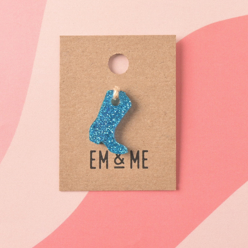 small, laser-cut acrylic mini charm shaped like a cowboy boot in vibrant blue glitter, secured to a brown kraft jewelry card branded with 'EM & ME.'