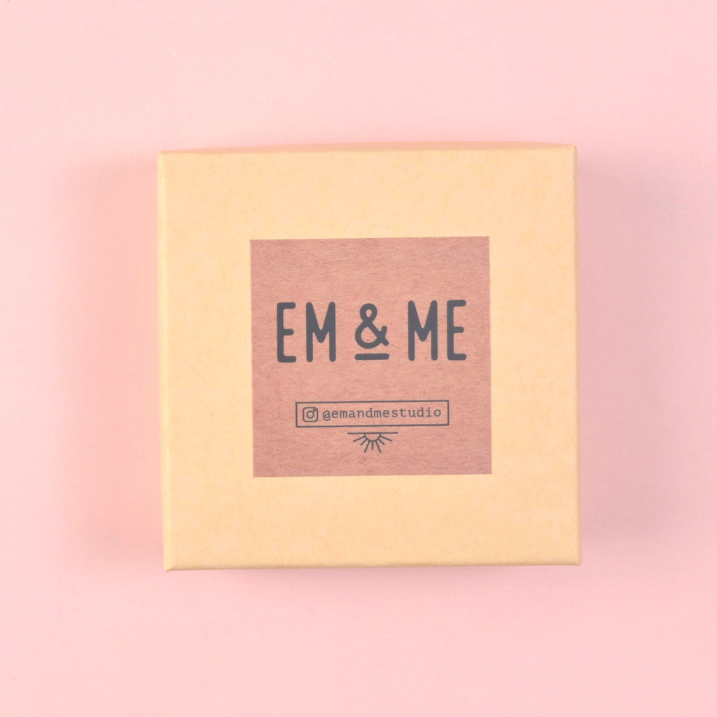 A closed, square, light tan kraft paper gift box with a pink sticker label on the lid featuring the black text logo 'EM & ME' and the social media handle '@emandmestudio'.