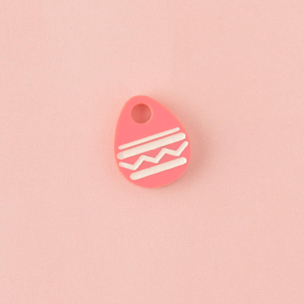 Close-up of a single, bright pink acrylic charm shaped like an Easter egg, decorated with white engraved zigzag and stripe patterns. The charm is displayed on a solid light pink background.