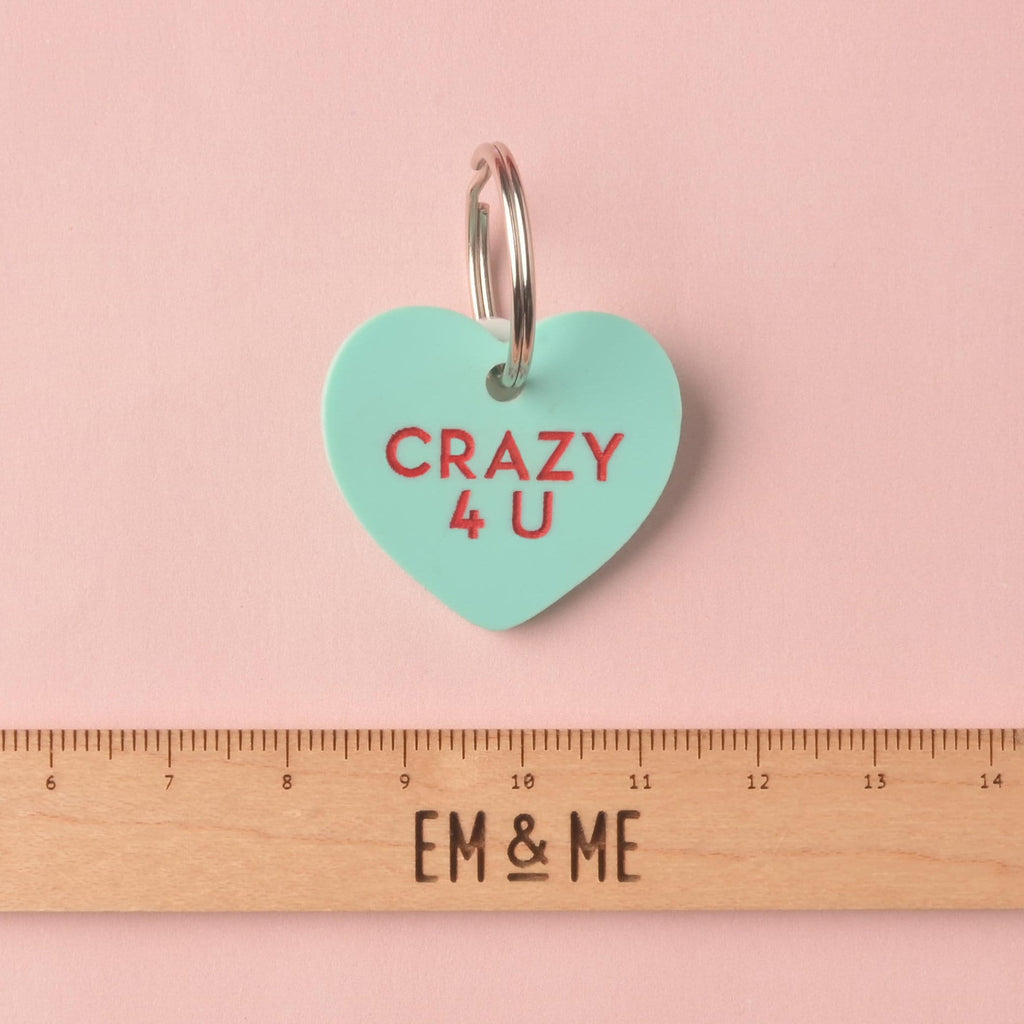 Close-up of a single heart-shaped acrylic pet ID tag in mint green, engraved with the red slogan 'CRAZY 4 U.' The tag is attached to a silver split ring and displayed above a wooden ruler marked with the brand EM & ME, on a pink background. Conversation Hearts, Valentines Heart Dog tag, Customize front and back, ID tag for cats and dogs, Personalized Pet Tag