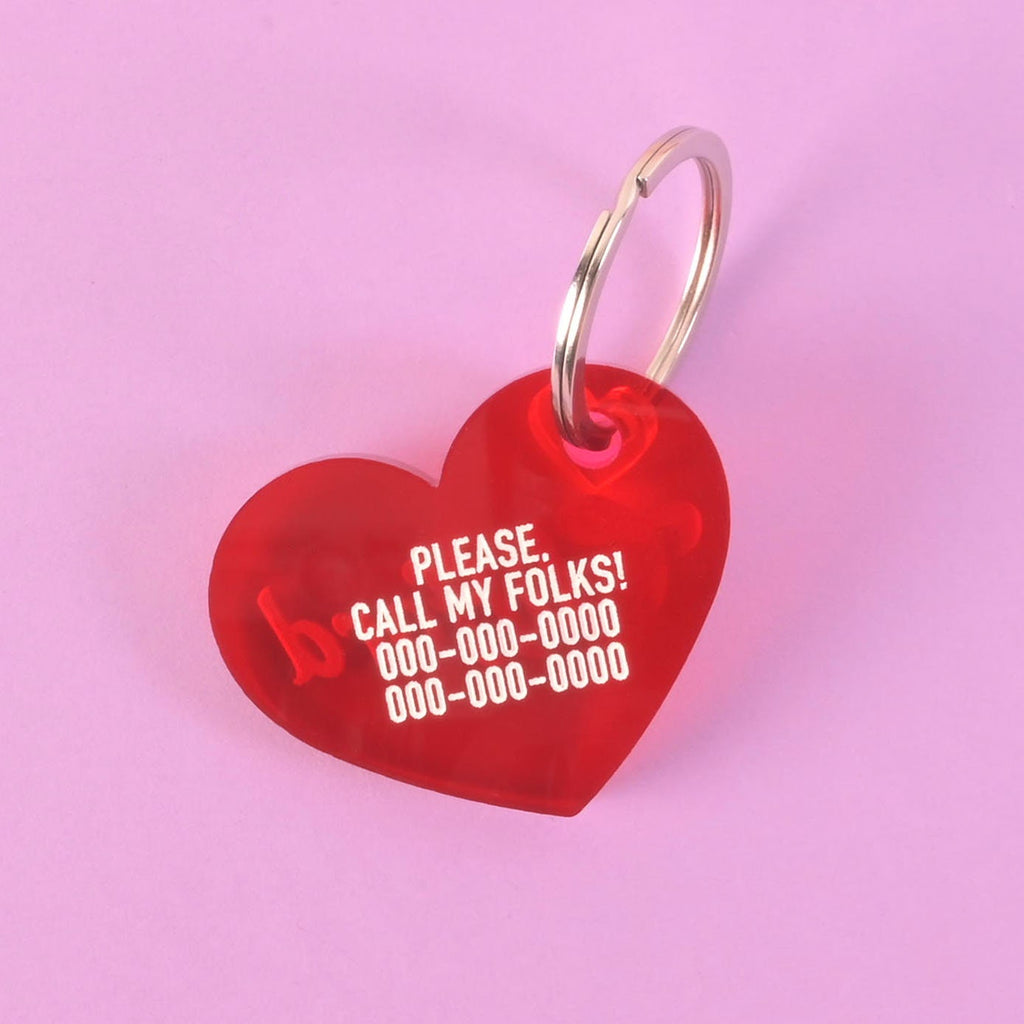 A personalized red heart-shaped pet tag that says "PLEASE. CALL MY FOLKS!" and has a placeholder phone number on it. The tag is attached to a silver ring, perfect for a collar, and is shown against a solid purple background.