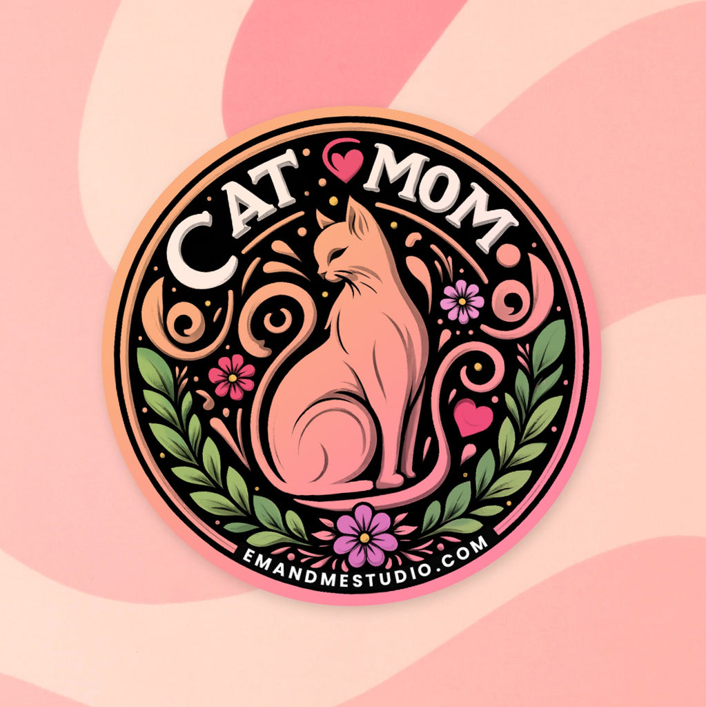 A close-up of a circular sticker featuring a pink cat in the center surrounded by a colorful floral design. The text at the top reads "CAT MOM" with "EM & ME STUDIO" at the bottom. The sticker is shown on a pink background with a wavy design.