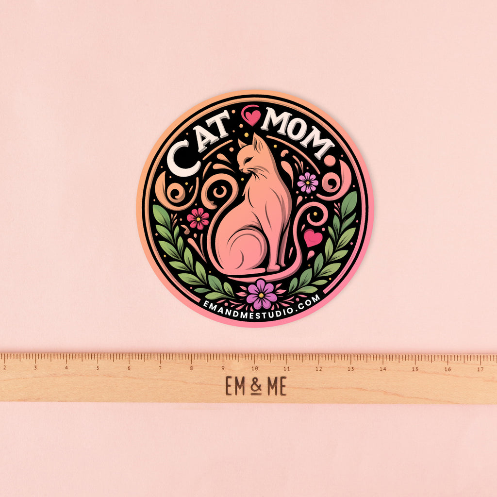 A white, circular sticker featuring a pink cat in the center surrounded by a colorful floral design. The text at the top reads "CAT MOM" with "EM & ME STUDIO" at the bottom. A wooden ruler with the "EM & ME" logo is placed below the sticker to show its size. The sticker is shown on a solid pink background.
