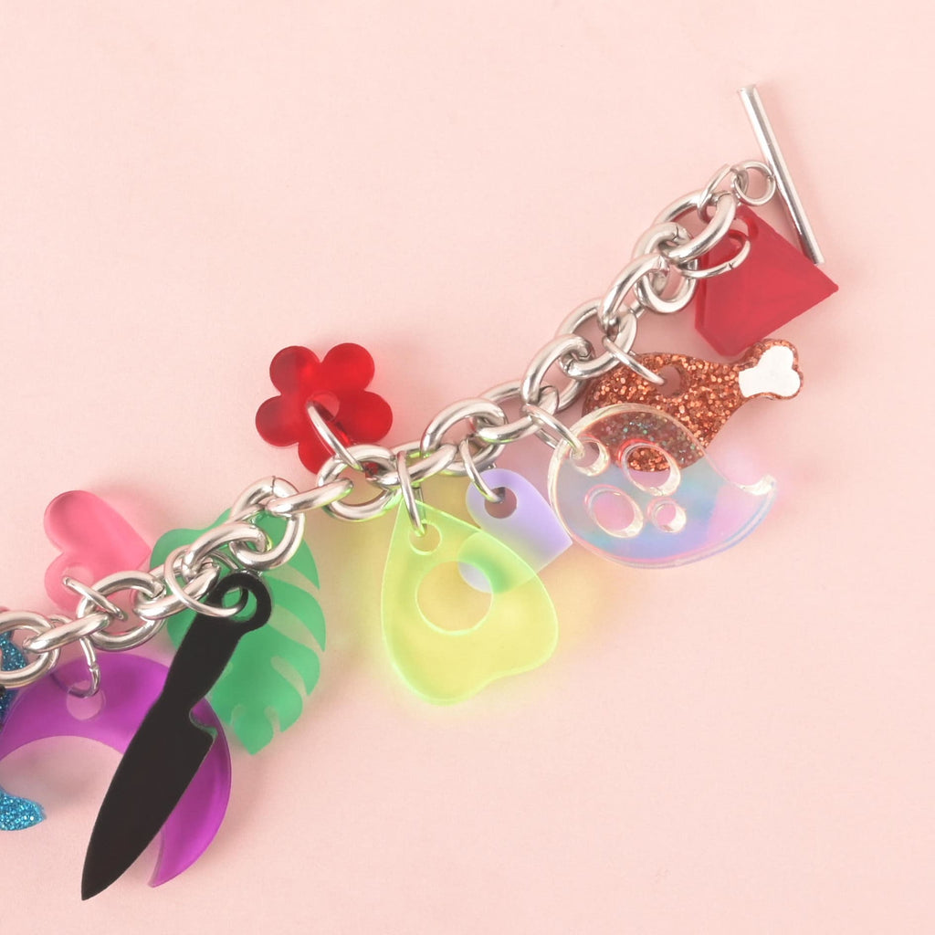 A silver charm bracelet with an assortment of spooky and quirky charms. The charms include a purple iridescent ghost, a black knife, a neon yellow planchette, a red drumstick, a pink seahorse, a red flower, a green leaf, a purple moon, a red crystal, and more. The bracelet is on a solid pink background.