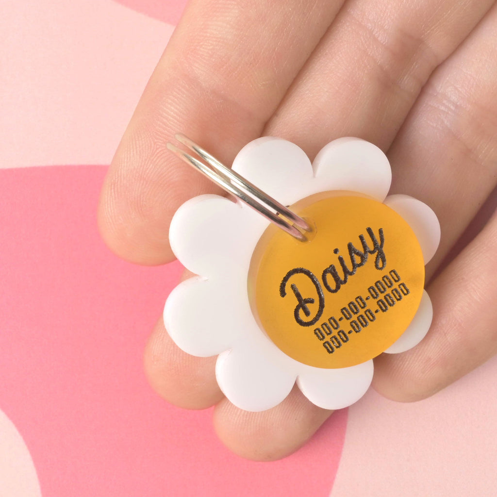 Hand holding a white flower-shaped pet tag with a yellow center engraved with the name "Daisy" and placeholder phone numbers, shown against a pink patterned background