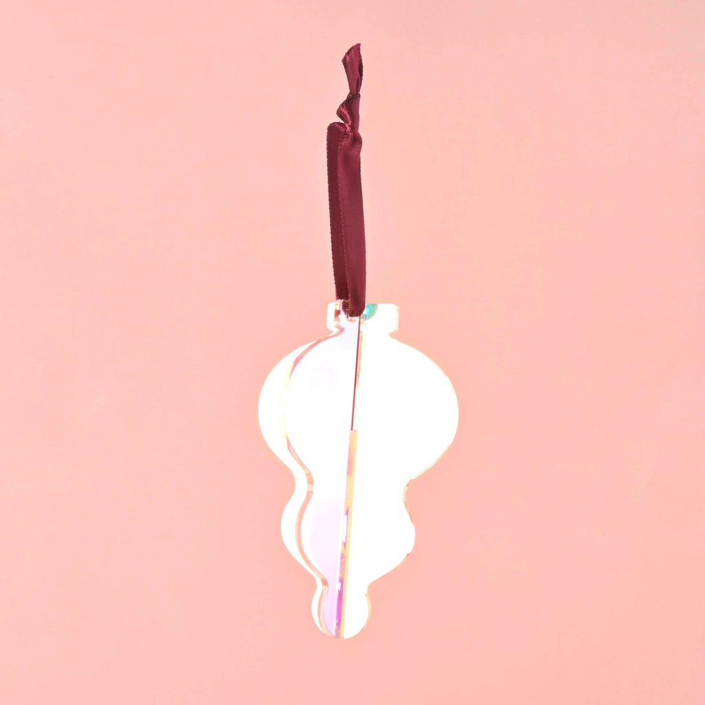 A single 3D interlocking acrylic Christmas ornament in a simple, curved, onion-bulb shape with an iridescent finish. It is hanging from a dark red/burgundy satin ribbon against a light pink background. Acrylic Icicle Christmas ornaments, bright colors and non-breakable, contemporary 70s inspired Holiday
