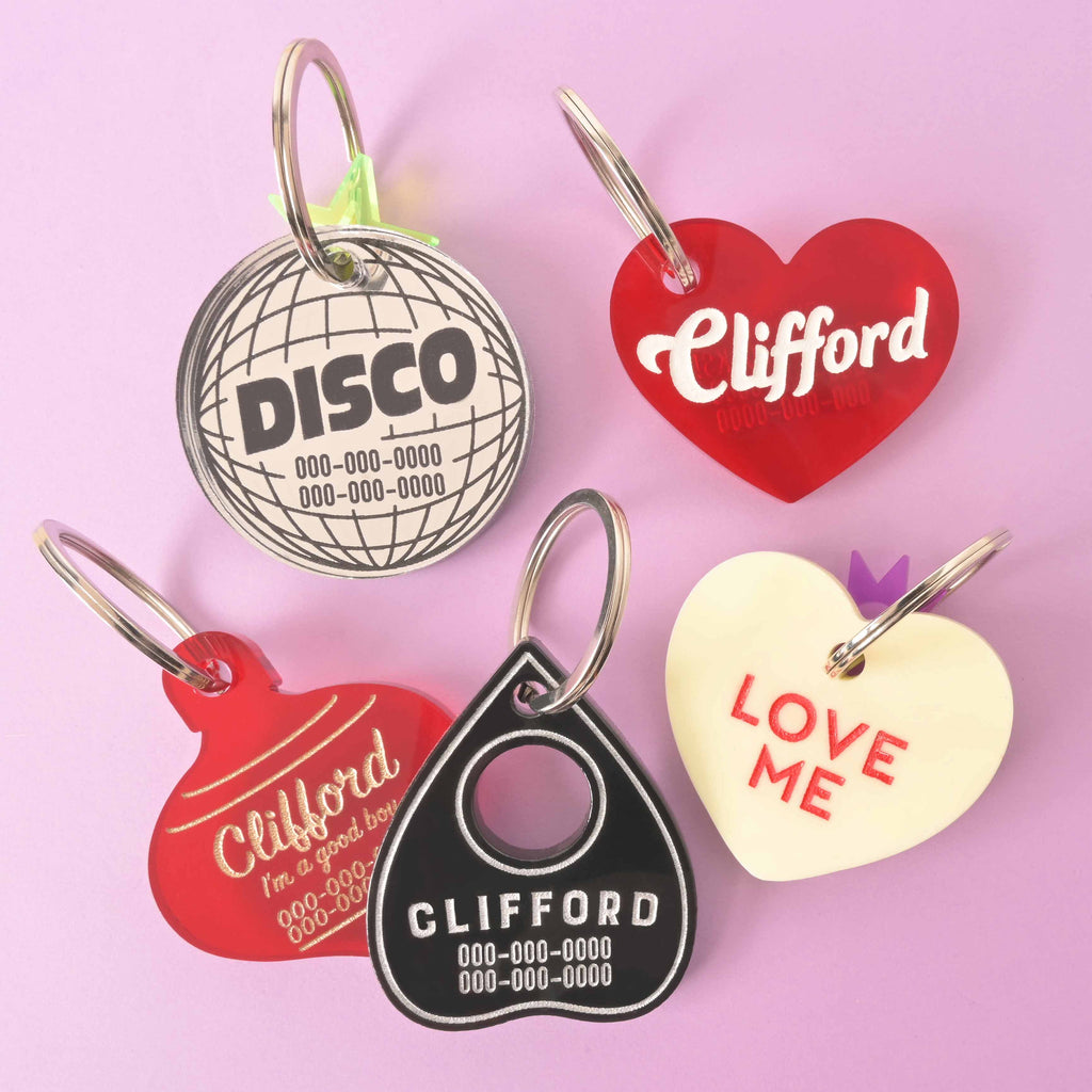 A set of five personalized pet tags in various shapes and colors scattered on a solid purple background. The tags include a silver disco ball, a red heart, a red "Clifford" ornament, a black Ouija planchette, and a white heart that says "LOVE ME."