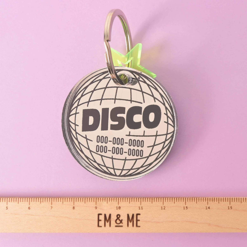 Large Disco Pet tag, Mirror Disco Personalized Pet Tag the Clifford Series, for big dog, extra strong ID Tag A personalized circular disco ball-shaped pet tag with the word "DISCO" and a placeholder phone number on it. A small, translucent yellow star charm is attached. A wooden ruler with the "EM & ME" logo is placed below the tag to show its size. The tag is displayed on a solid purple background.