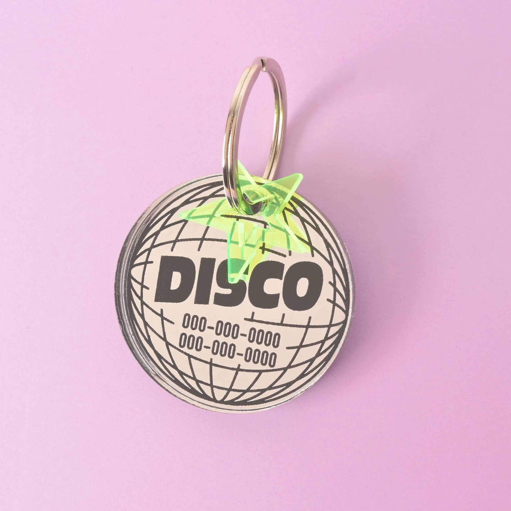 A personalized circular disco ball-shaped pet tag with the word "DISCO" and a placeholder phone number on it. A small, translucent yellow star charm is attached. The tag is displayed on a solid purple background.