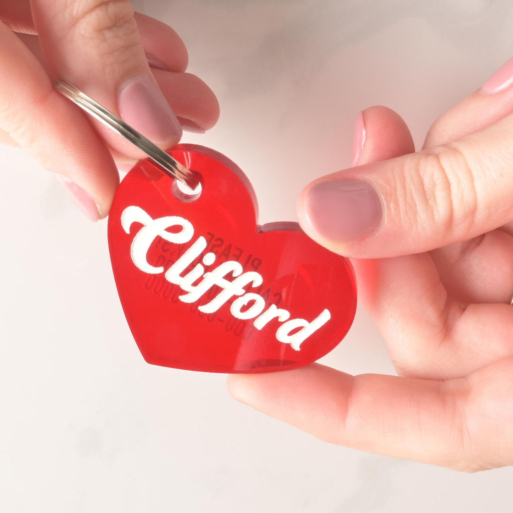 A person's hands hold a personalized red heart-shaped pet tag. The tag has the name "Clifford" and a placeholder phone number on it. It's attached to a silver ring, perfect for a collar, and is shown against a white marble surface.