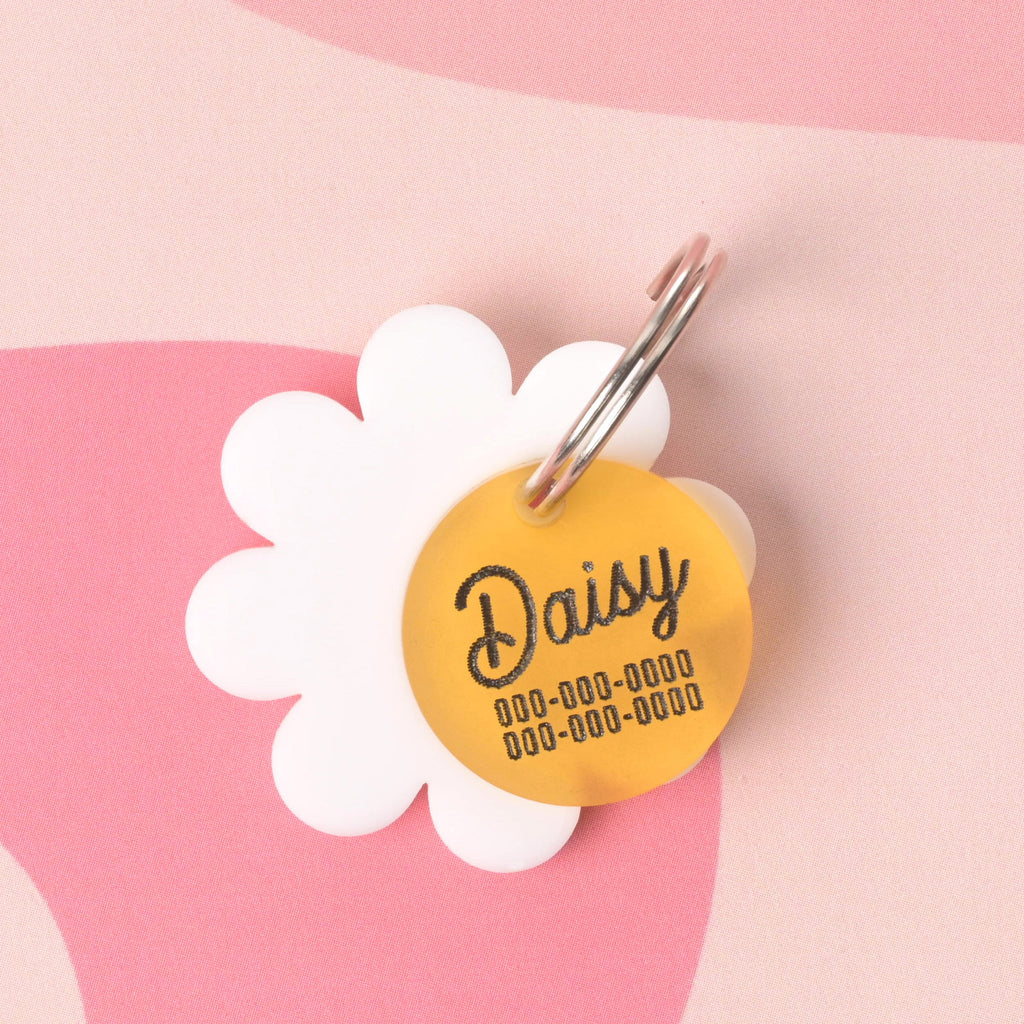 White flower-shaped pet ID tag with a yellow center engraved with the name "Daisy" and placeholder phone numbers, displayed on a pink wavy checkerboard background