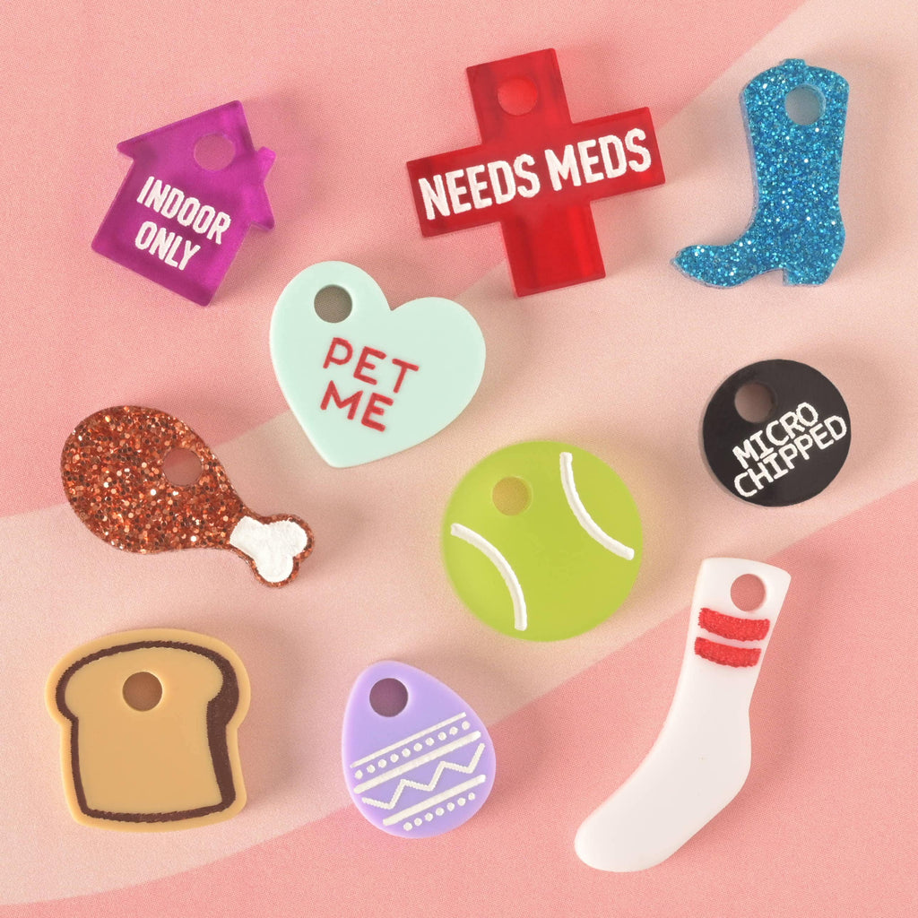 A scattered collection of colorful pet charms and tags. Designs include a red cross that says "NEEDS MEDS," a mint green heart that says "PET ME," a black circle that says "MICROCHIPPED," a purple house that says "INDOOR ONLY," a blue glitter cowboy boot, a glittery brown chicken leg, a green tennis ball, a piece of toast, a purple Easter egg, and a white tube sock. The items are displayed on a pink and white background.