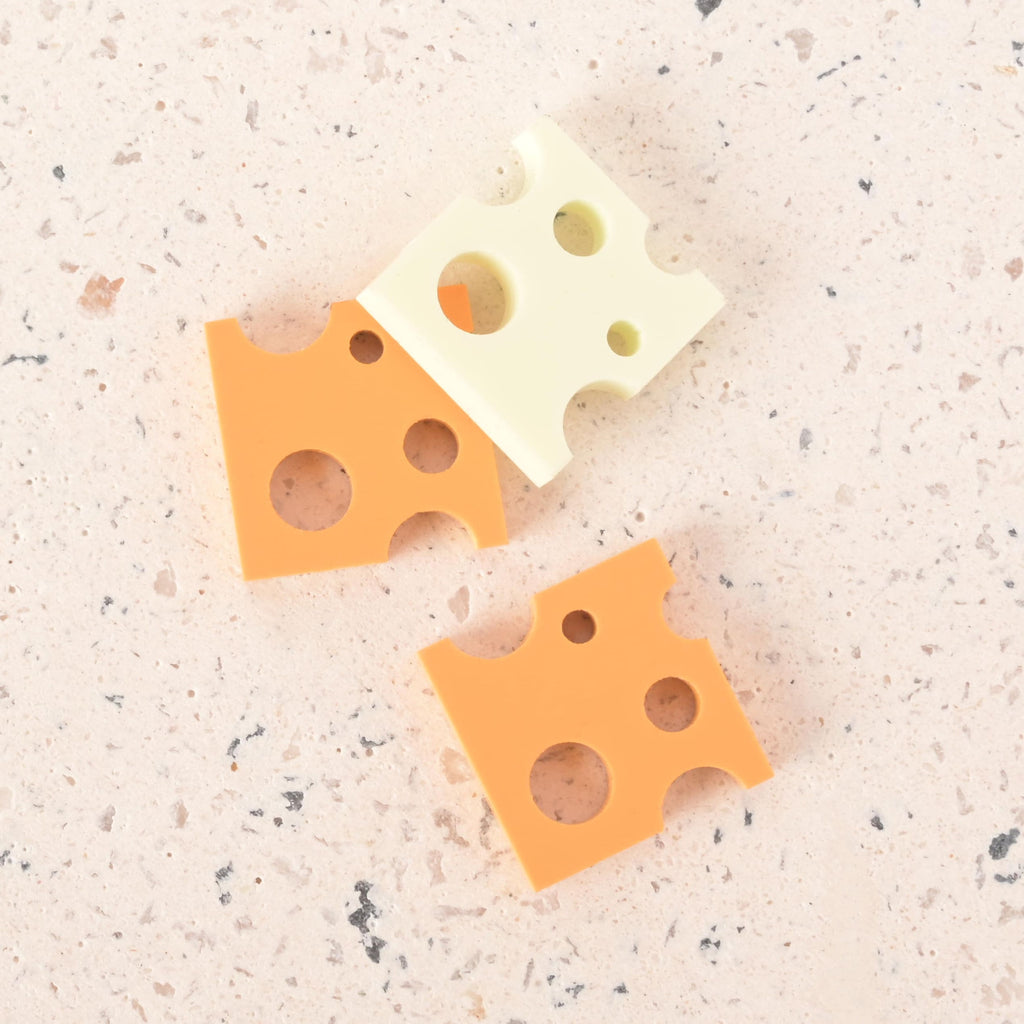 A group of three cheese-shaped charms. One is a light yellow-white color, while the other two are a more vibrant orange-yellow. The charms have various-sized holes and are shown on a light-colored, speckled surface.