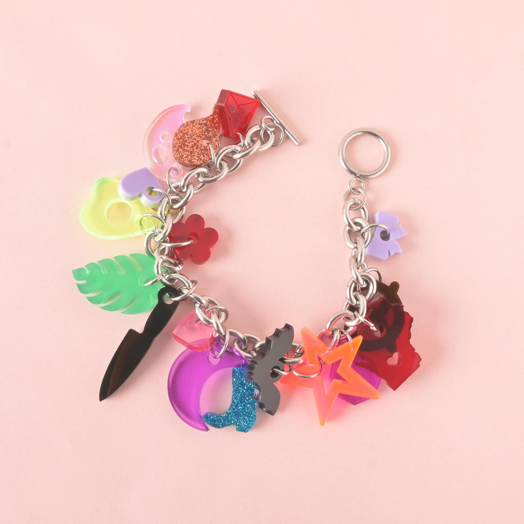 A silver charm bracelet with an assortment of colorful charms. The charms include a purple crescent moon with a blue glittery boot, a black knife, a green monstera leaf, a red flower, a neon pink and orange star, a red Wisconsin-shaped charm with a heart, and a translucent ghost, among others. The bracelet is on a solid pink background.