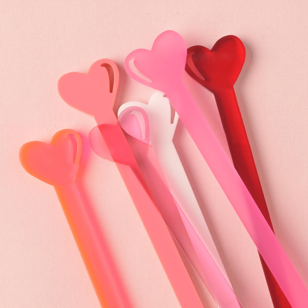 A close-up, fanned cluster of heart-tipped acrylic stir sticks in varying shades of red, bright pink, light pink, and white. They are displayed diagonally against a solid light pink background.