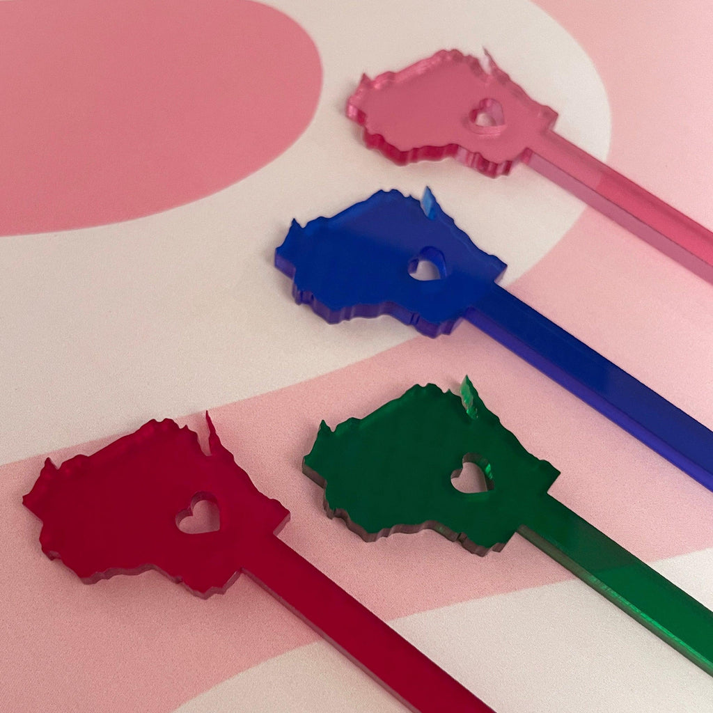 Four acrylic Wisconsin-shaped drink stir sticks with heart cutouts in pink, blue, green, and red, displayed diagonally on a pink and white abstract background.