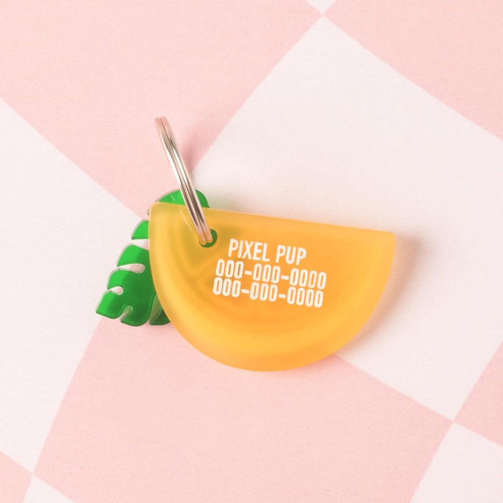A personalized, semi-translucent yellow/orange acrylic pet ID tag shaped like a citrus slice, engraved with the name 'PIXEL PUP' and three lines of phone numbers. A small green tropical leaf charm is attached to the tag ring.