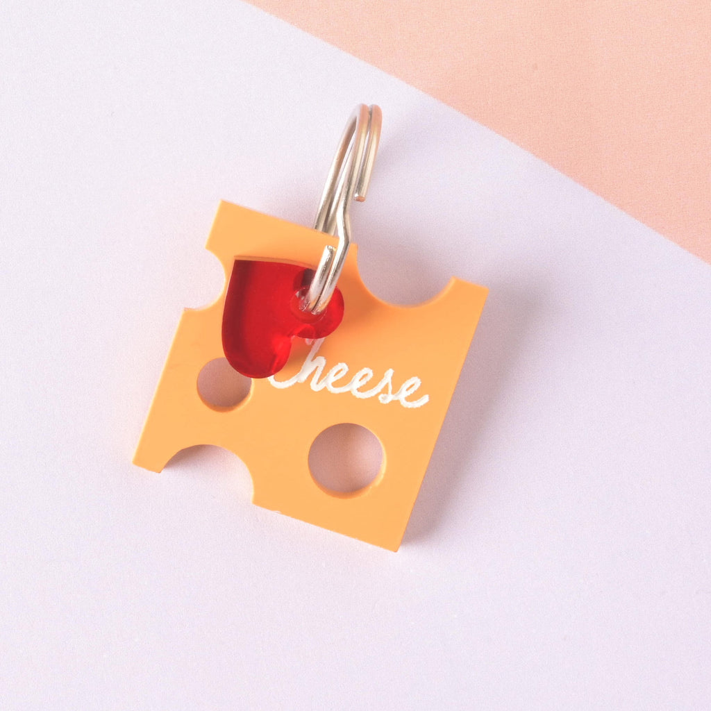 A custom pet ID tag shaped like a slice of Swiss cheese (orange acrylic) with "holes" cut out. The tag is engraved with the word "Cheese" and accented with a small translucent red heart charm, attached to a gold split ring.