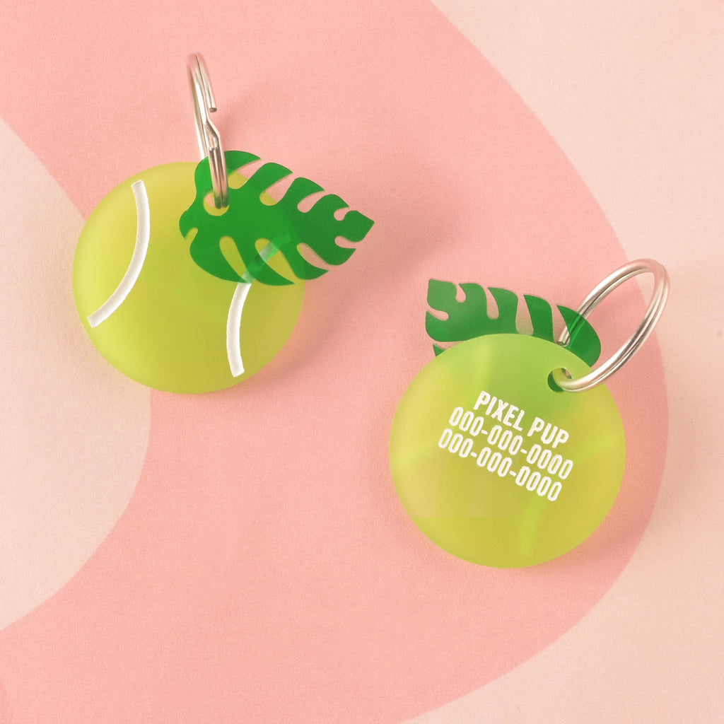 A personalized pet ID tag shaped like a tennis ball in fluorescent neon green acrylic. The back is engraved with the name 'PIXEL PUP' and contact information, and the tag is accented with a dark green tropical Monstera leaf charm. Tennis Ball Pet Tag, Personalized Pet tag, Cat or Dog ID Tag, summer ball tag