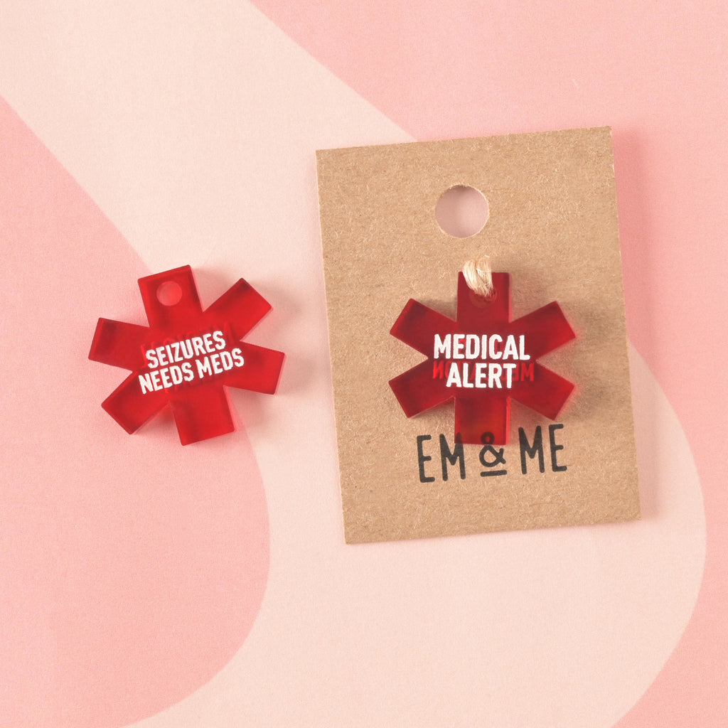 Two red translucent acrylic medical alert charms shaped like the Star of Life. One is engraved with "MEDICAL ALERT" and is shown on a branded kraft card. The other is engraved with "SEIZURES NEEDS MEDS."