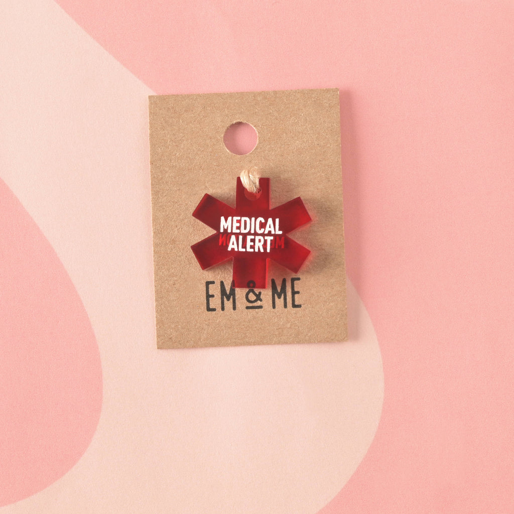 A translucent red acrylic medical alert charm shaped like the Star of Life. The charm is engraved with "MEDICAL ALERT" and secured with twine to a branded 'EM & ME' kraft card.