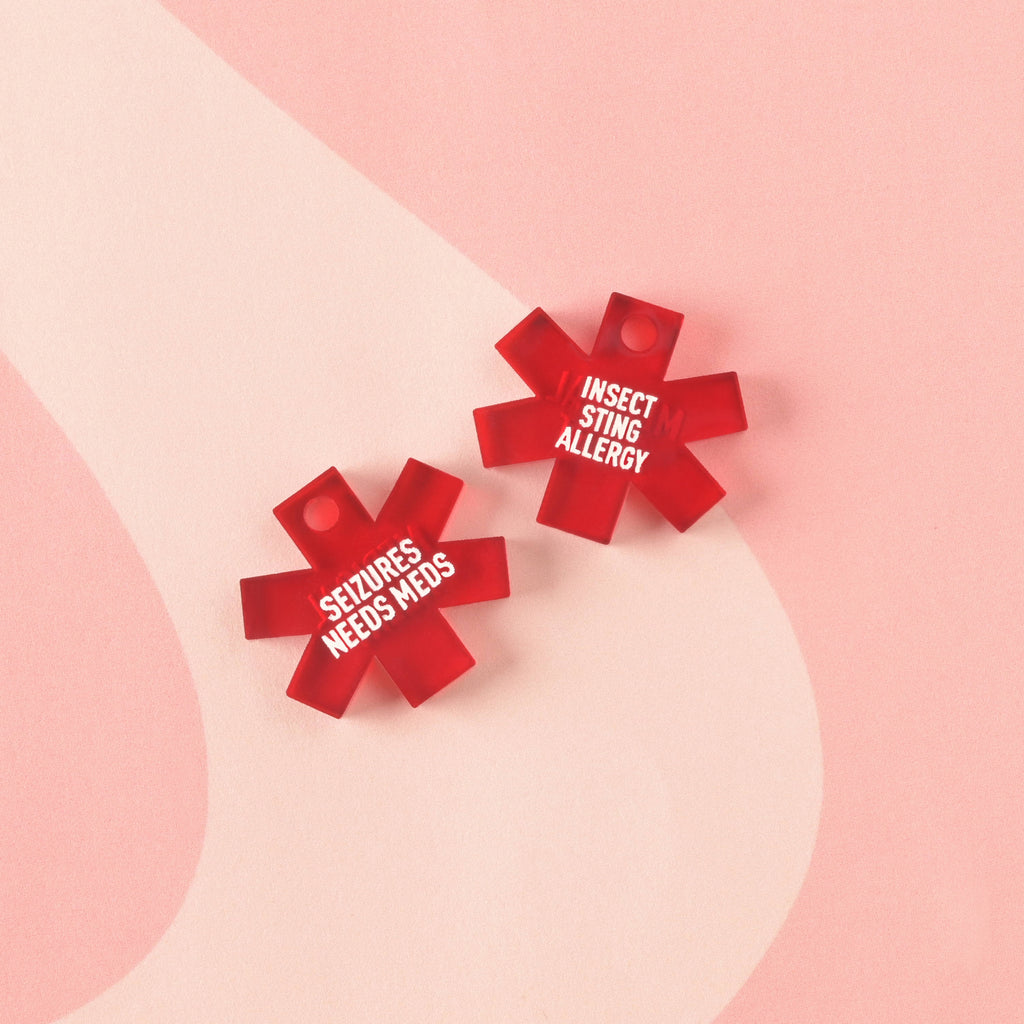 Two translucent red acrylic medical alert charms shaped like the Star of Life. One is engraved with "SEIZURES NEEDS MEDS" and the other with "INSECT STING ALLERGY" in white text.