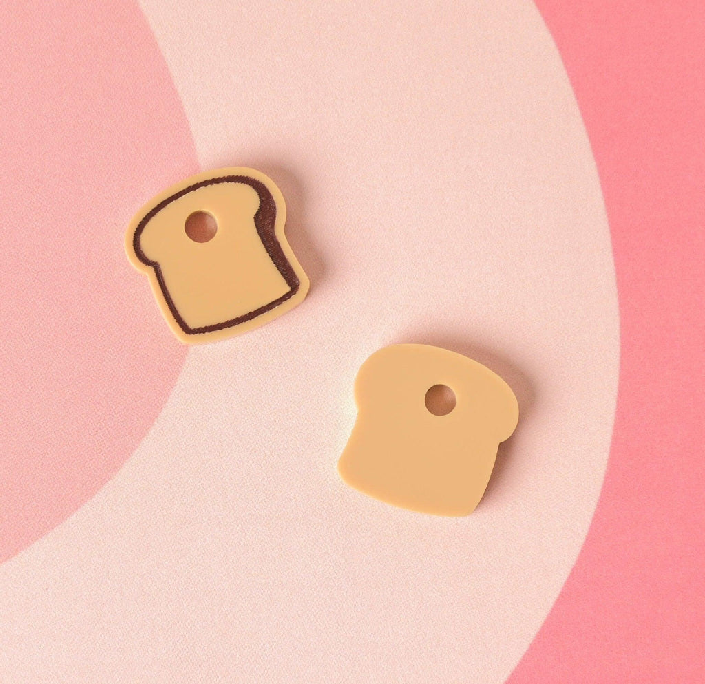 Two small, light brown/tan acrylic charms shaped like slices of toast. The charm on the left has a dark brown crust outline, while the charm on the right is solid light brown. They are displayed on a pink patterned background. Bread Mini Charm, Tiny Toast Accessory for Pet ID Tag, Cats and Dogs Add-on Charm
