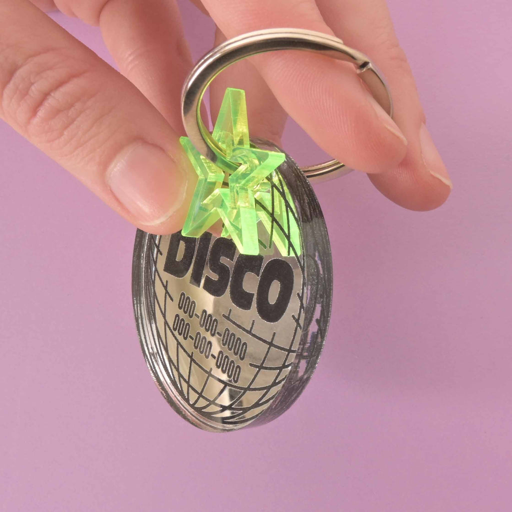 A hand holding a disco ball-shaped pet ID tag that says "DISCO" and has two phone numbers. The tag also has a green star charm.