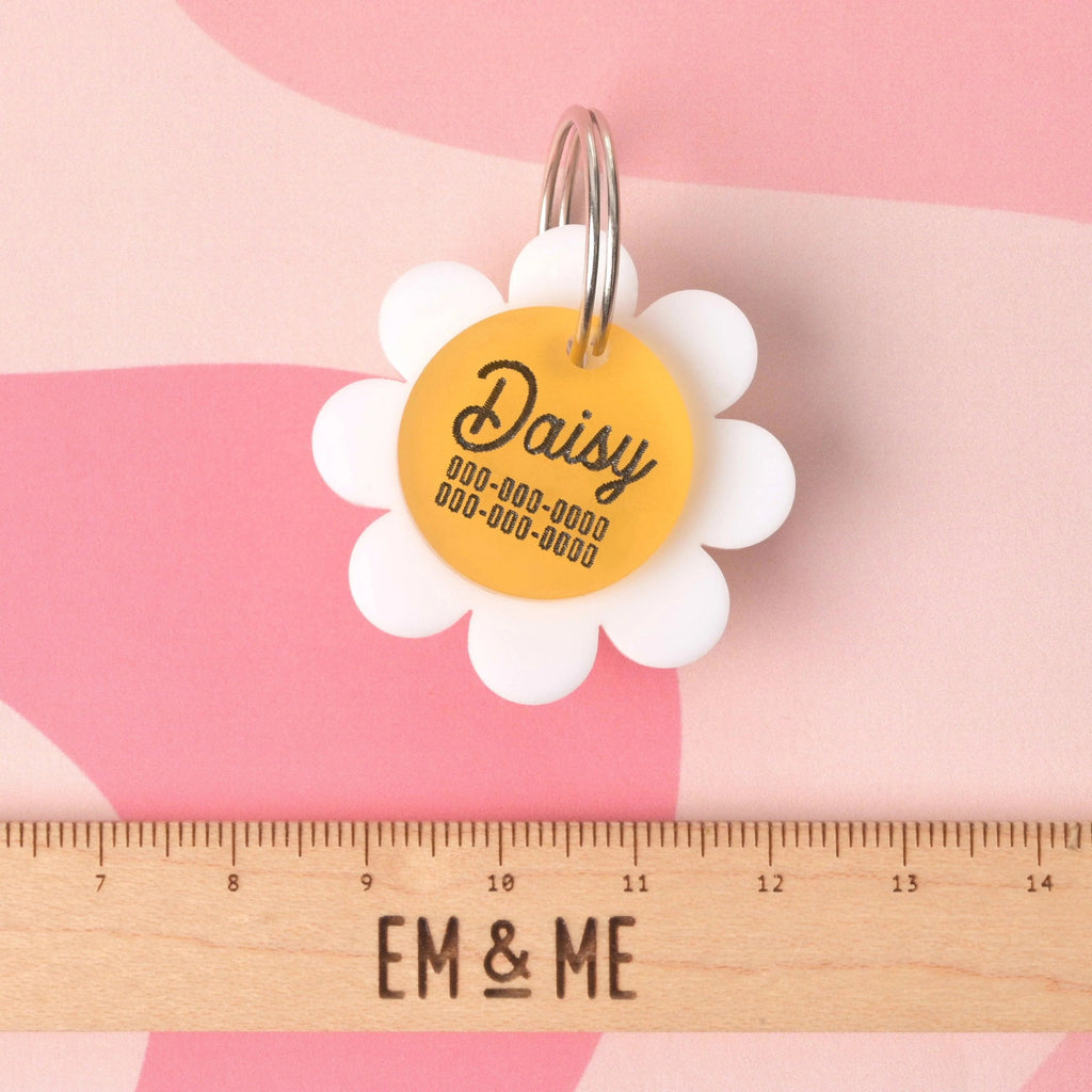 Custom daisy-shaped acrylic pet ID tag with the name Daisy and engraved contact numbers, shown above a wooden EM & ME Studio ruler for size reference, on a pink abstract background
