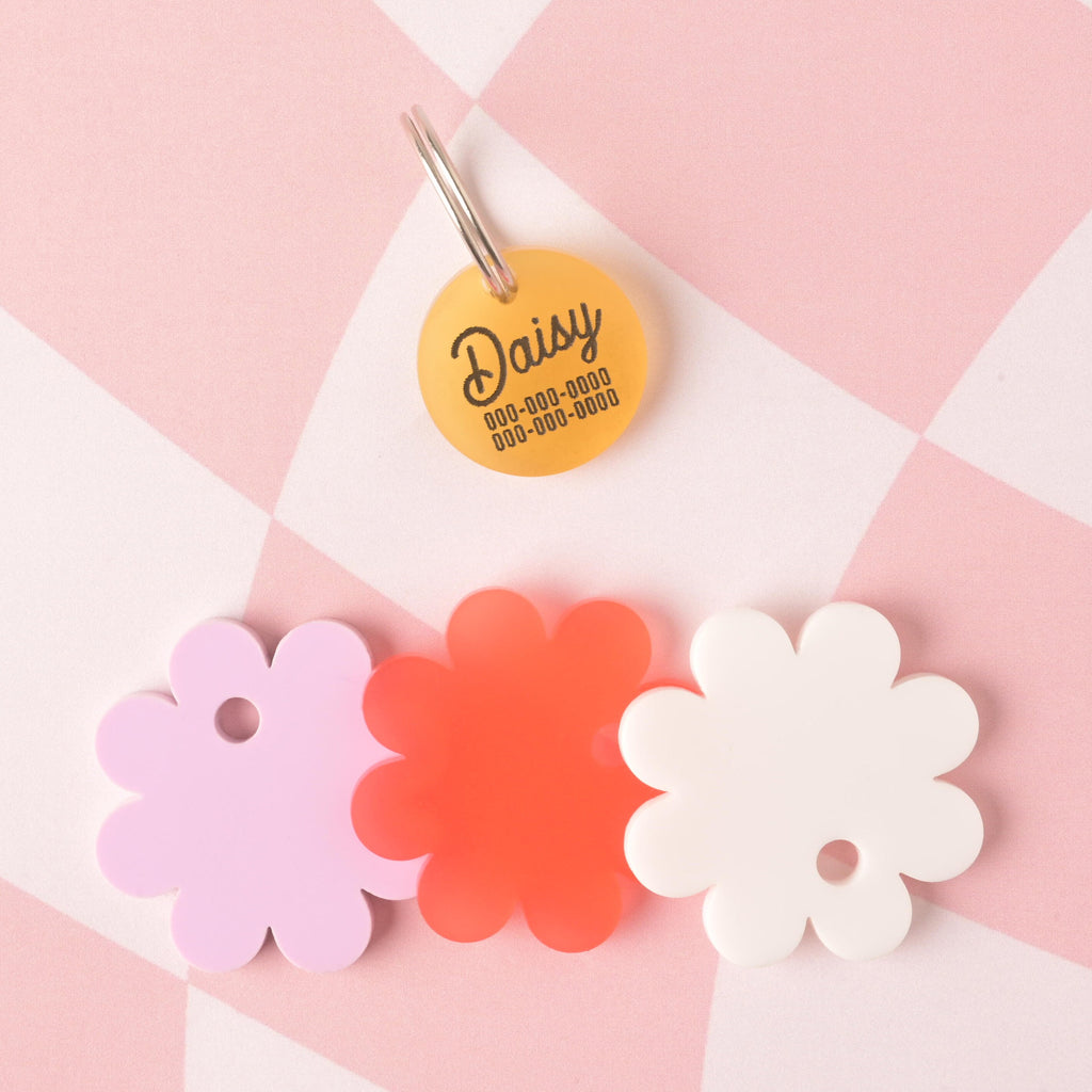 Yellow circular pet ID tag with the name "Daisy" shown above three flower-shaped acrylic backings in lavender, poppy red, and white, arranged on a pink checkered background