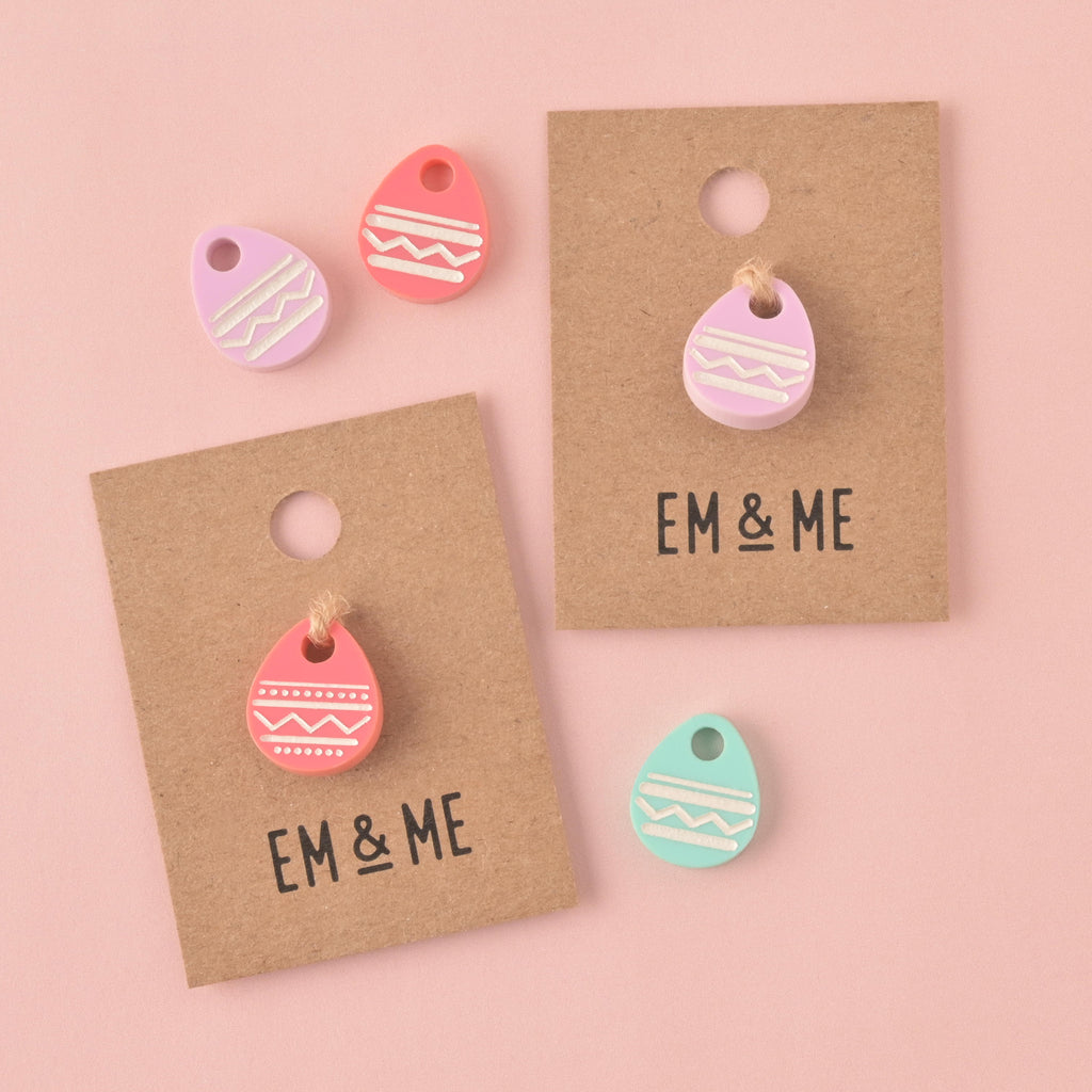 A collection of five pastel Easter egg-shaped acrylic pet charms (pink, light purple, and mint green). Two charms are attached to EM & ME branded kraft paper backing cards, and three are loose, all against a light pink background.