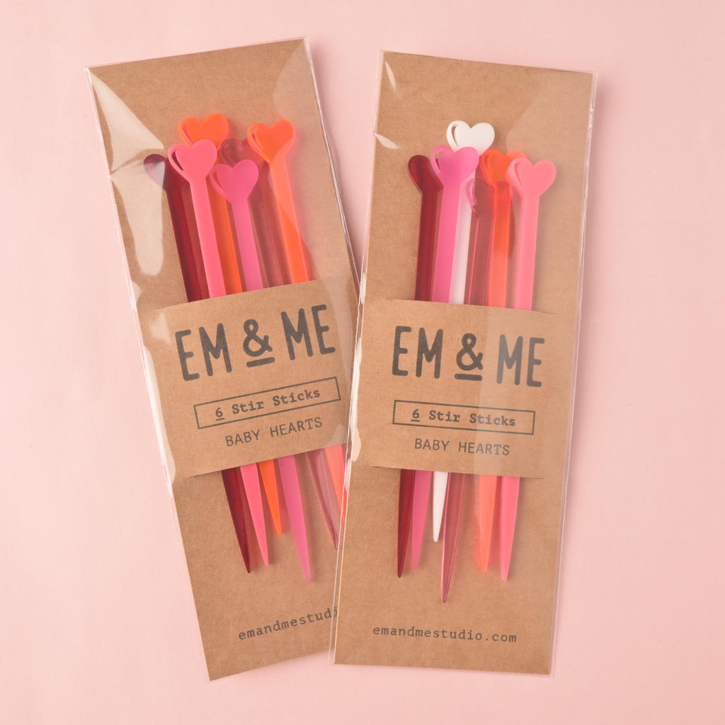 Two packaged sets of EM & ME 'Baby Hearts' acrylic stir sticks. Each package contains 6 heart-tipped sticks in various shades of pink, red, and orange, displayed in clear sleeves with a kraft paper backing card.